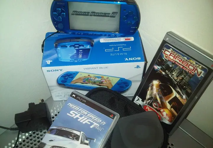 Niebieska konsola PSP z grą Ridge Racer 2, pudełko z konsolą i grami Need for Speed Shift i Carbon. Klasyczne stare gry na telefon.